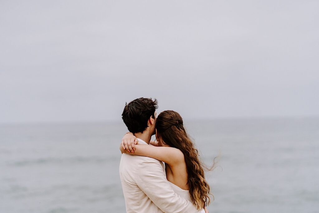 A bride and groom embrace one another and look out at the ocean while on the cliffs in San Diego while celebrating their destination elopement