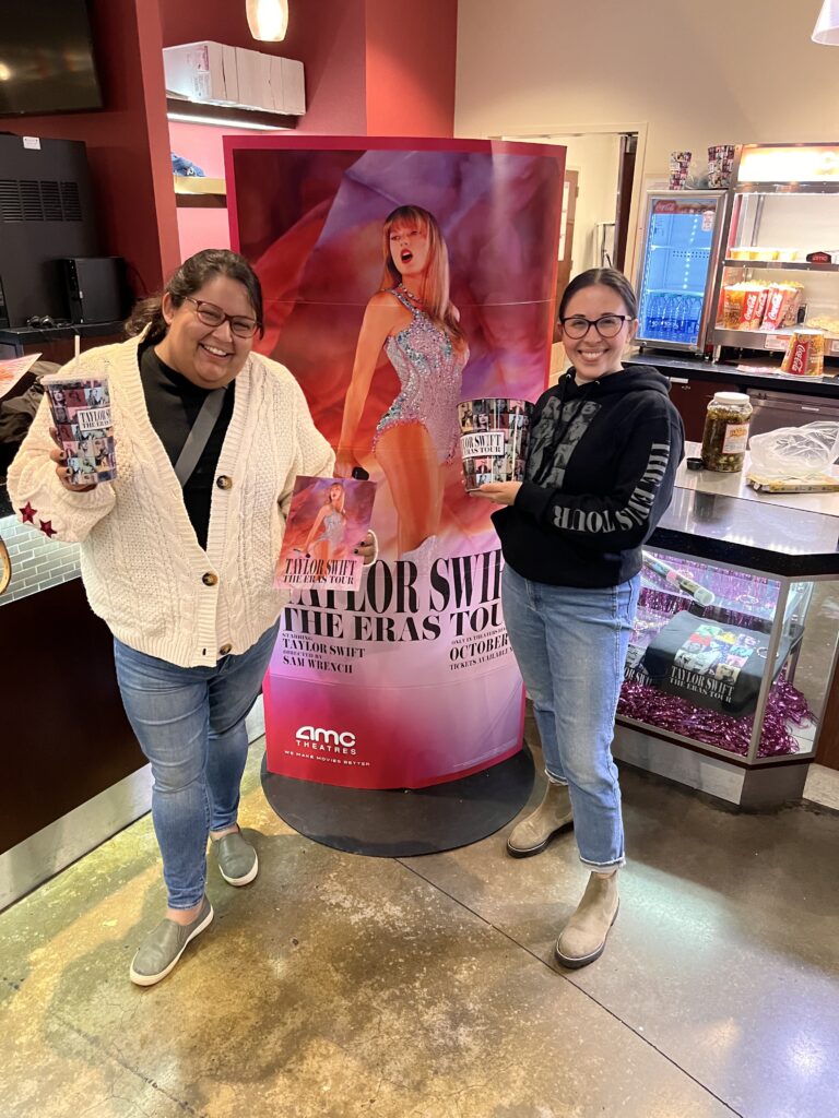 Two women smile at the camera while standing next to an ad for Taylor Swift's movie and holding popcorn