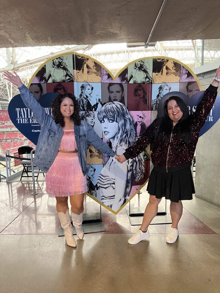 Two women hold hands and smile at the camera, between them is a massive heart collage of images of Taylor Swift set up at a concert
