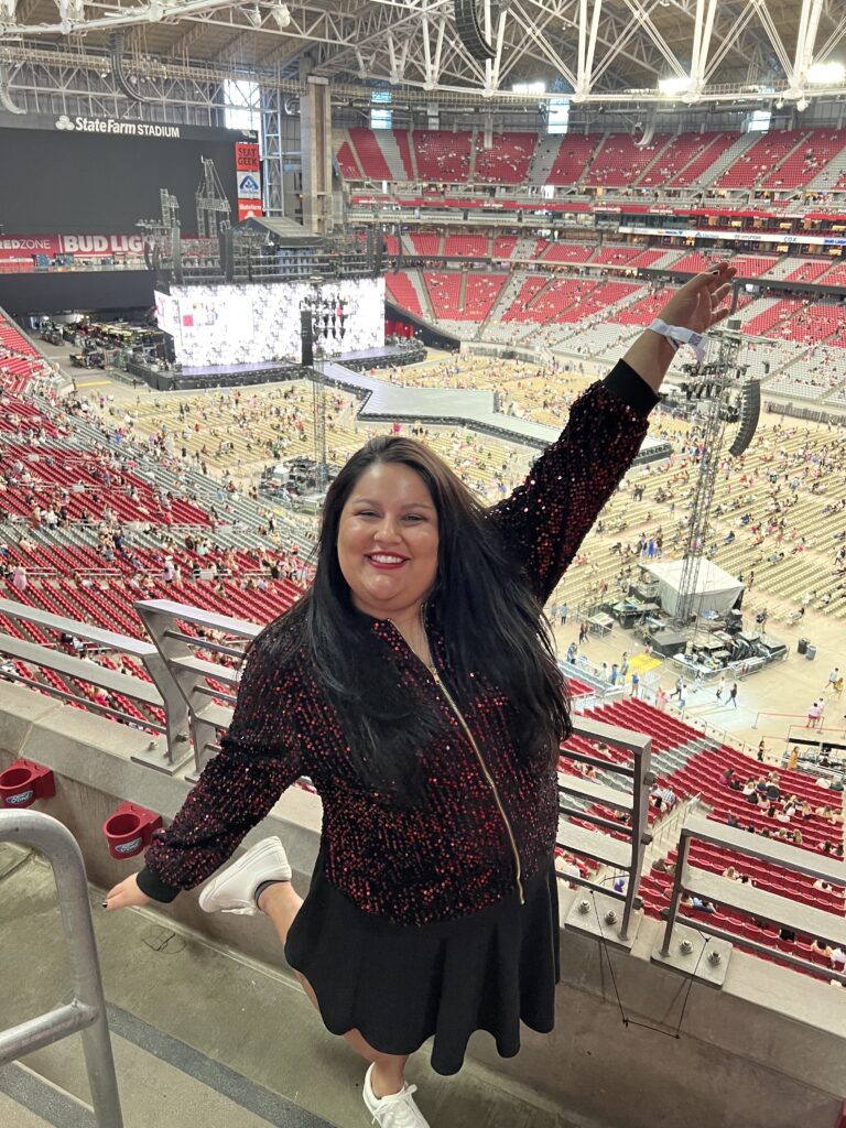 A woman (JNA Visuals) smiles at the camera while lifting an arm and a leg in the air while in a coliseum, behind her is the stage of a concert