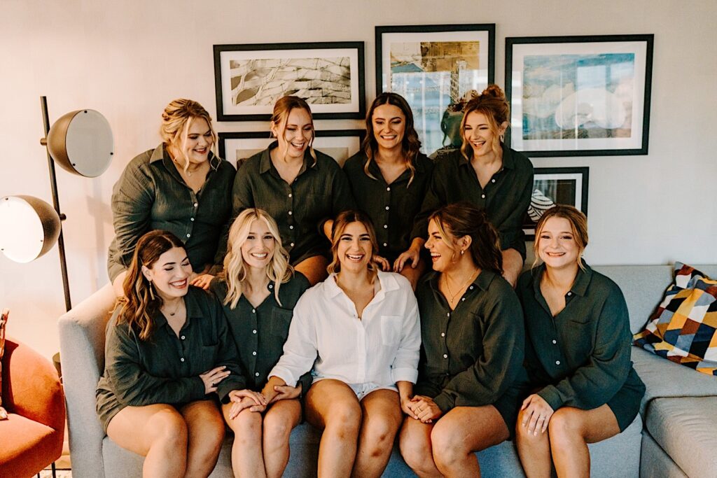 A bride sits on a couch surrounded by her 8 bridesmaids as they all smile before getting ready for the wedding