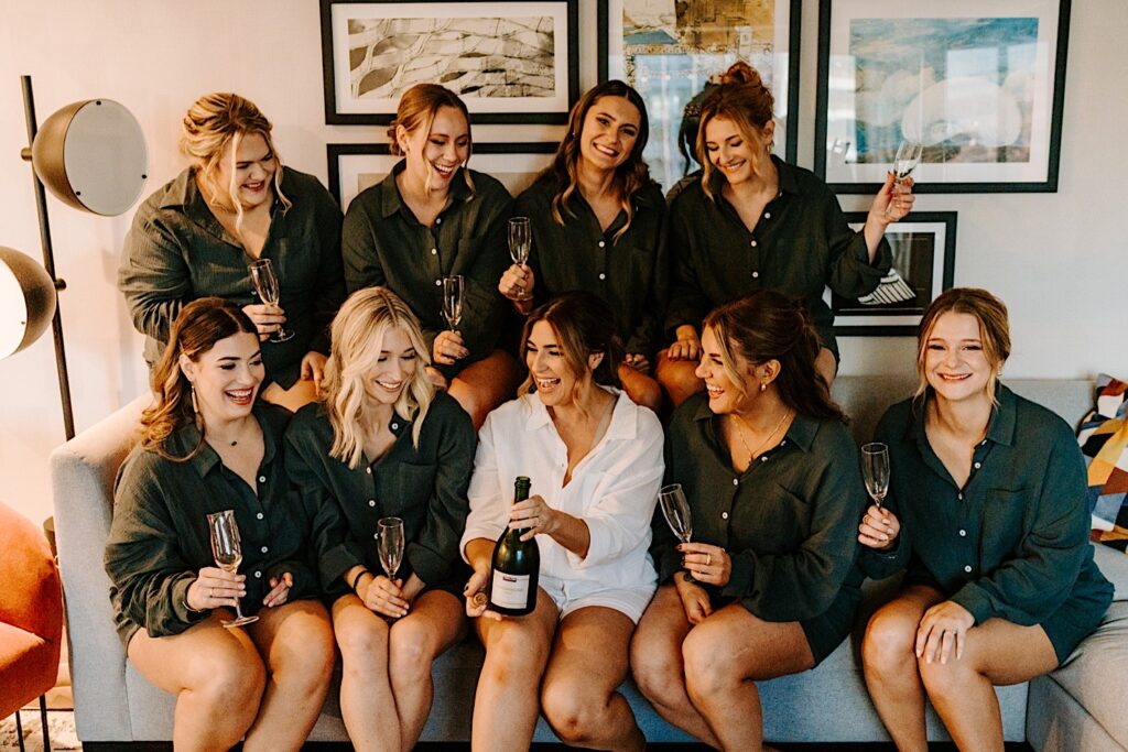 A bride sits on a couch surrounded by her 8 bridesmaids, the bride has a bottle of champagne and each bridesmaid has a glass