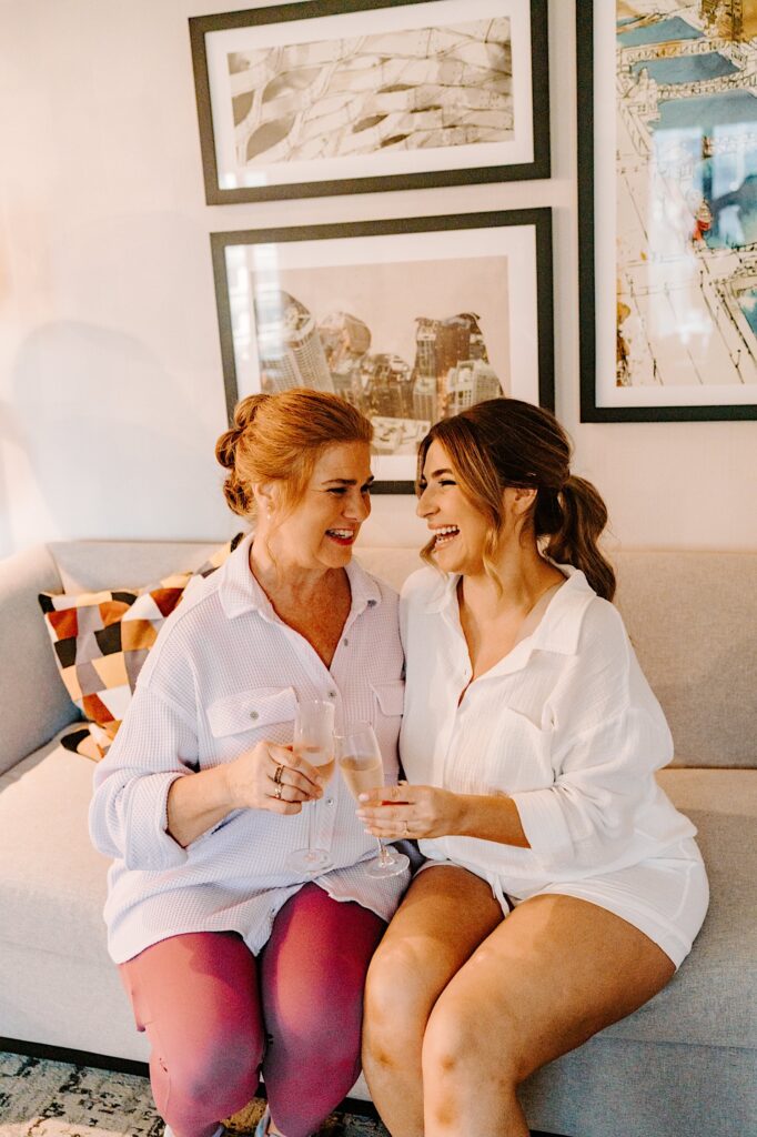 A bride sits on a couch next to her mother as the two laugh while sipping champagne before getting ready for the wedding