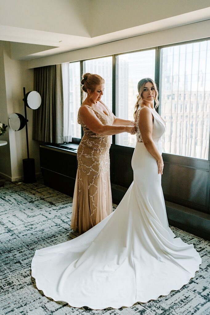 A bride stands in front of a window and looks over her shoulder as her mother buttons up the back of her dress for her