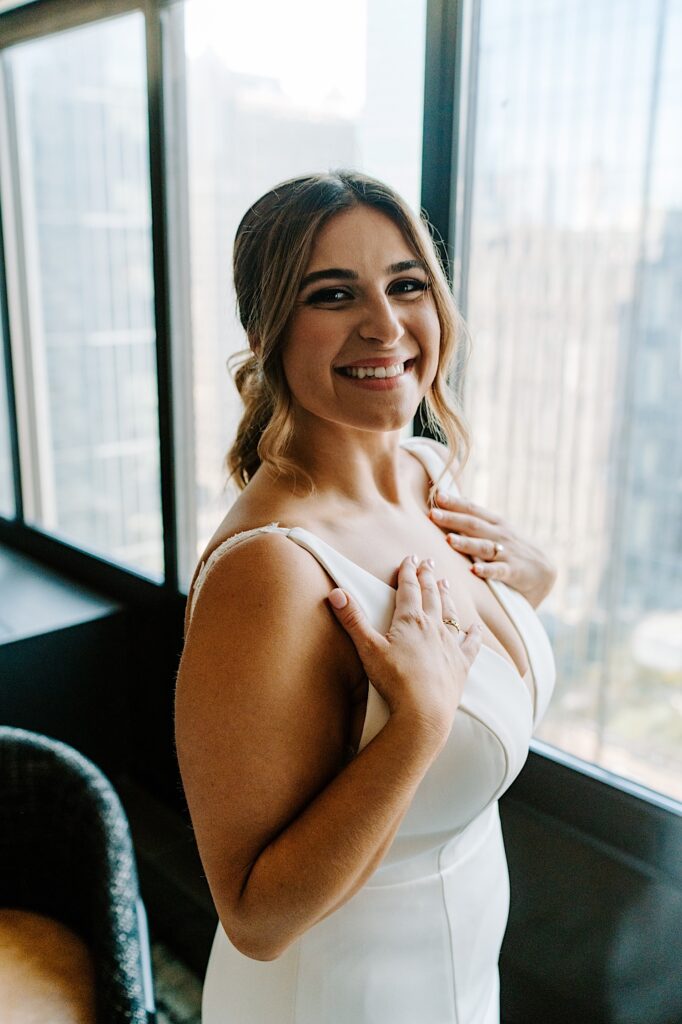 A bride stands in front of a window and smiles at the camera while in her wedding dress