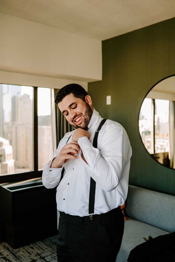 A groom smiles while looking down as he buttons up his shirt sleeve in his hotel room