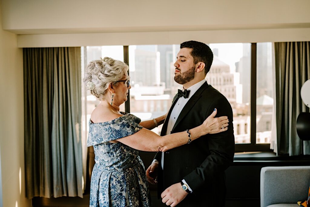 A mother looks over her son who is dressed for his wedding day and standing in his hotel room