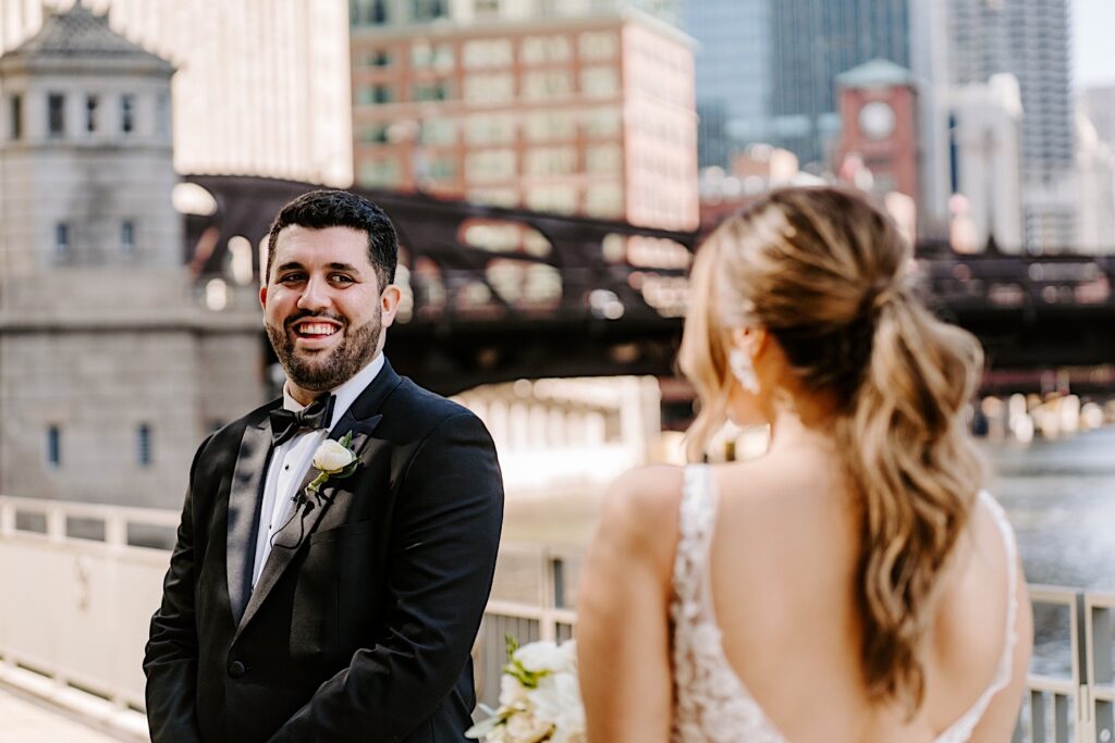 A groom smiles as he sees the bride in her wedding dress for the first time while the two stand next to the Chicago River outside the Voco Hotel