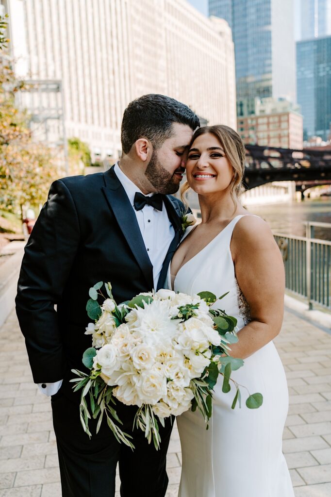 A bride smiles at the camera while the groom goes in to kiss her cheek while they stand next to the Chicago River