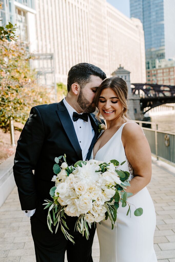 A bride smiles as she looks down at her bouquet while the groom kisses her cheek while the two stand next to the Chicago River