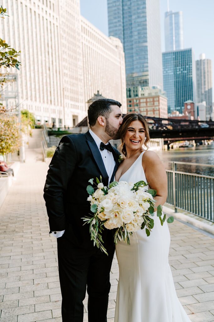 A bride smiles as the groom kisses her on the head while they stand on a walkway next to the Chicago River