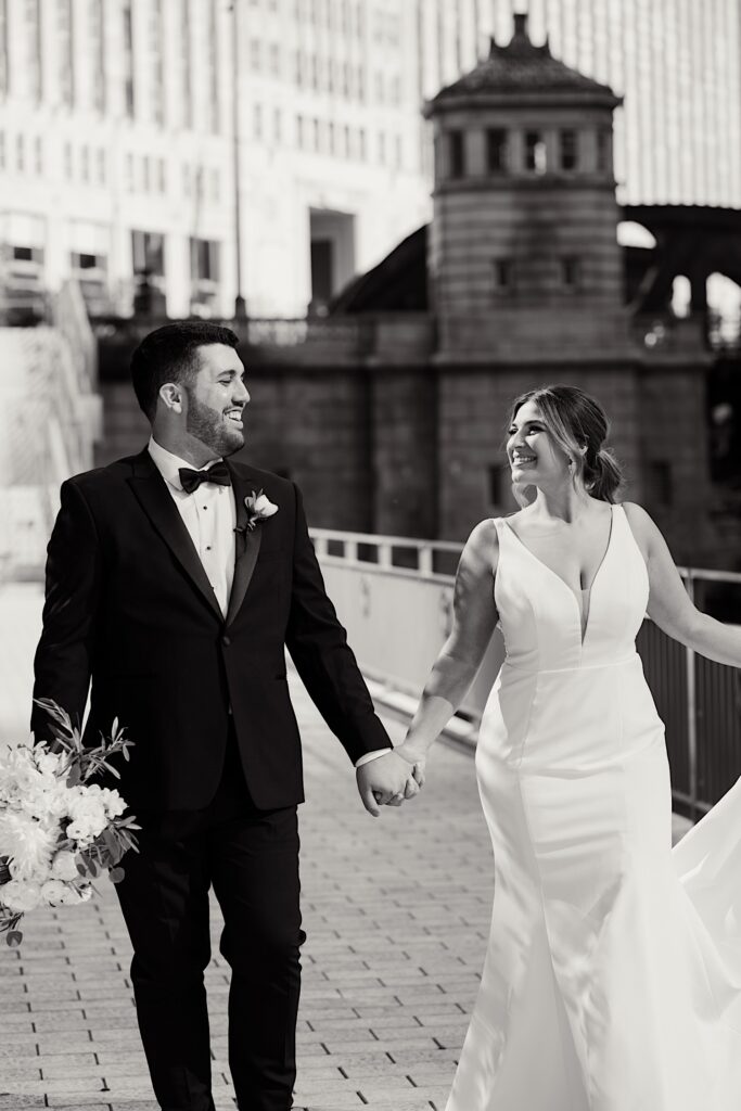 Black and white photo of a bride and groom walking hand in hand along the Chicago Riverwalk and smiling at one another