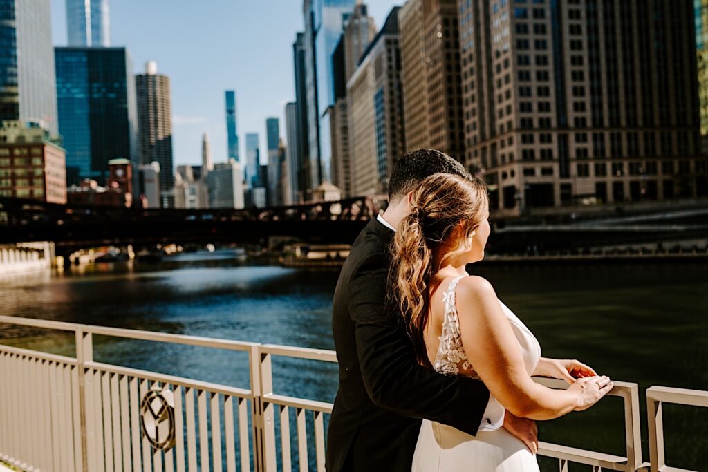 A groom wraps his arm around the bride as the two look over a railing at the Chicago River while outside their wedding venue, the Voco Hotel
