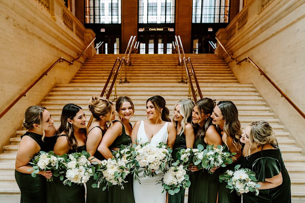 A bride stands with her 8 bridesmaids in front of a staircase inside Chicago's Union Station as the bridesmaids all smile towards the bride