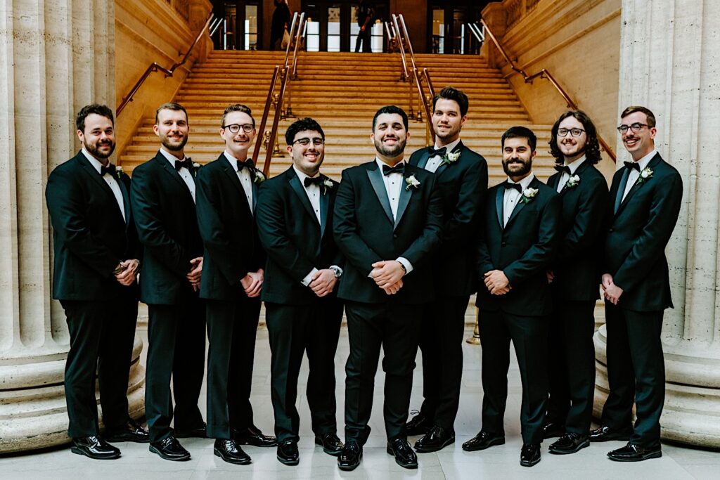 A groom stands with his 8 groomsmen inside of Chicago's Union Station for a group portrait in front of a staircase