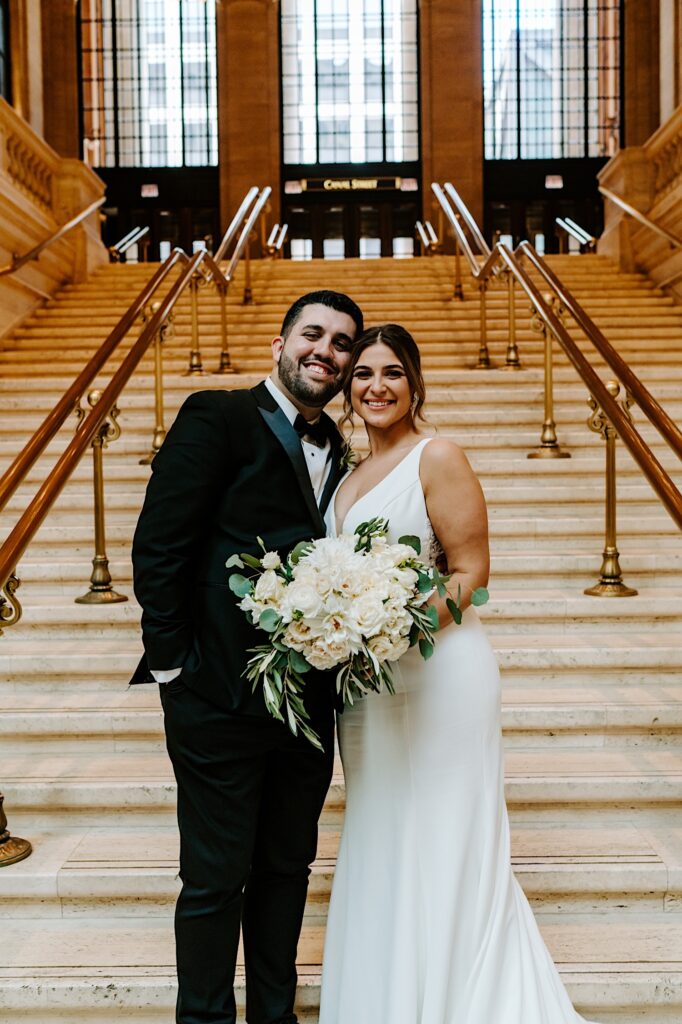 A bride and groom stand on a staircase in Chicago' Union Station for a portrait photo as they smile at the camera