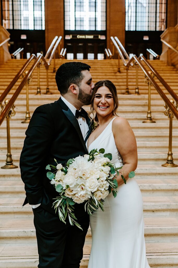 A bride smiles at the camera while the groom kisses her head while the two stand on a staircase inside of Chicago's Union Station