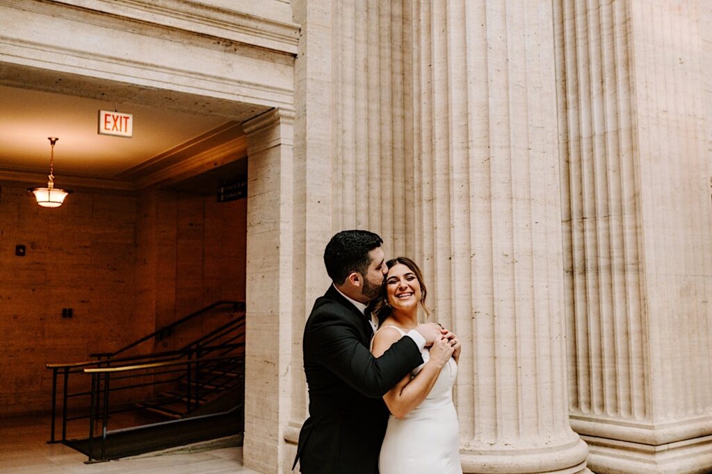 A bride smiles at the camera while the groom hugs her from behind and kisses her head while the two are inside of Chicago's Union Station