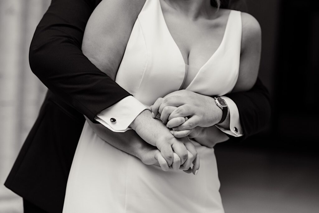 Black and white photo of a bride being hugged from behind by the groom, the focal point of the photo is the couple's hands intertwined