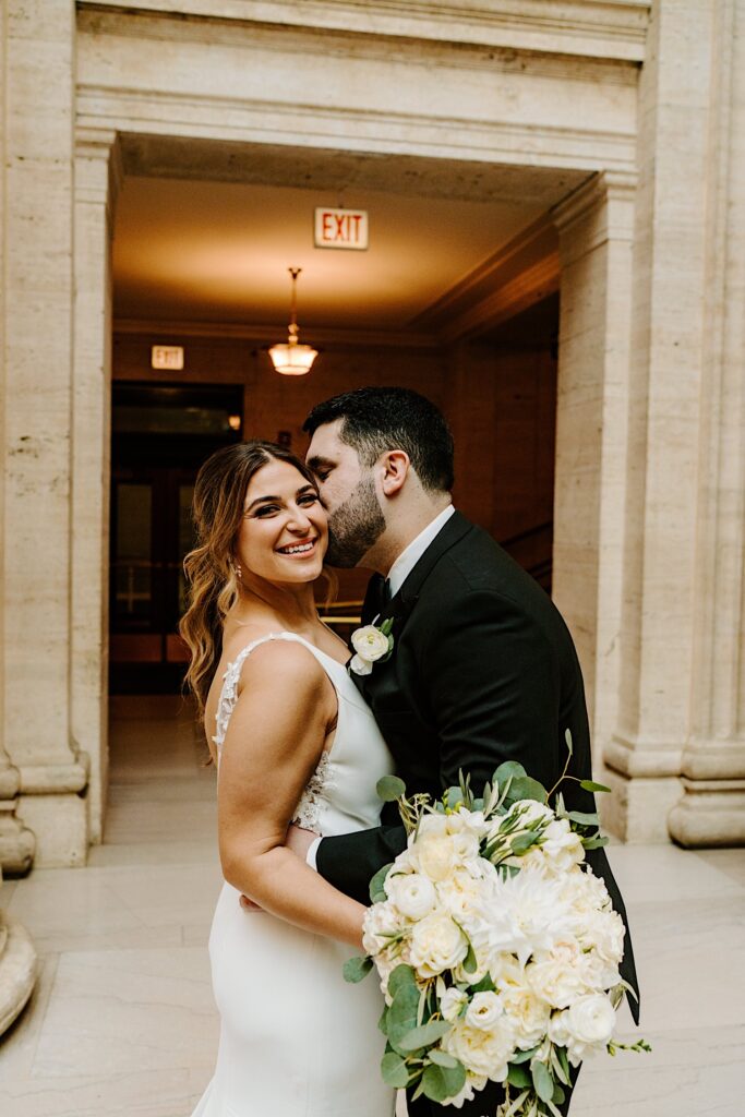 A bride smiles at the camera while the groom hugs her and kisses her cheek as the two stand inside Chicago's Union Station