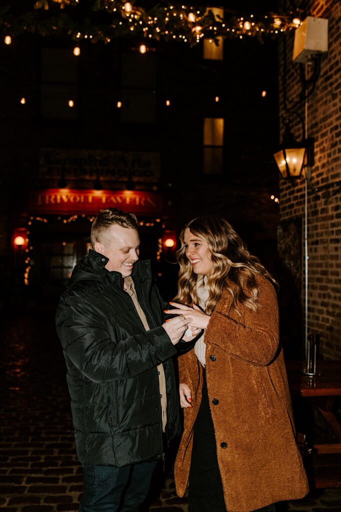 A couple smiles while standing outside of the Trivoli Tavern in Chicago as the man puts an engagement ring on the woman's finger