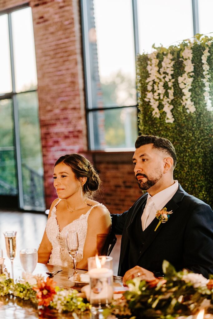 A bride smiles while the groom puts his arm around her as the two sit together at their sweetheart table