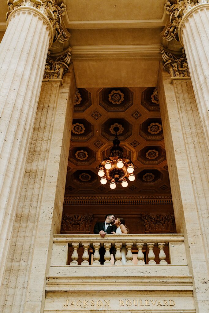 A bride and groom smile at one another and lean in for a kiss while looking out over a balcony inside of Chicago's Union Station
