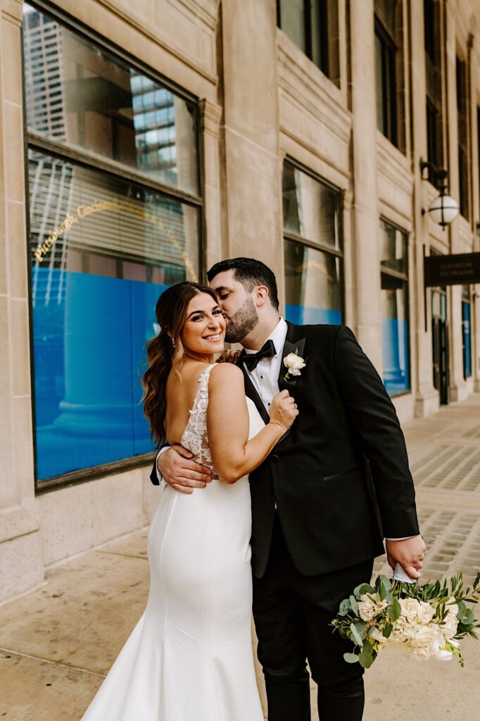 A bride smiles over her shoulder at the camera as the groom holds her and kisses her cheek outside of Chicago's Union Station