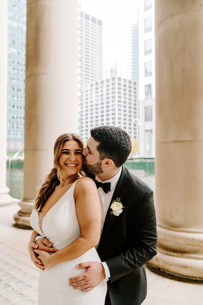 A bride smiles at the camera while the groom stands behind her and kisses her cheek as the two stand outside of Chicago's Union Station