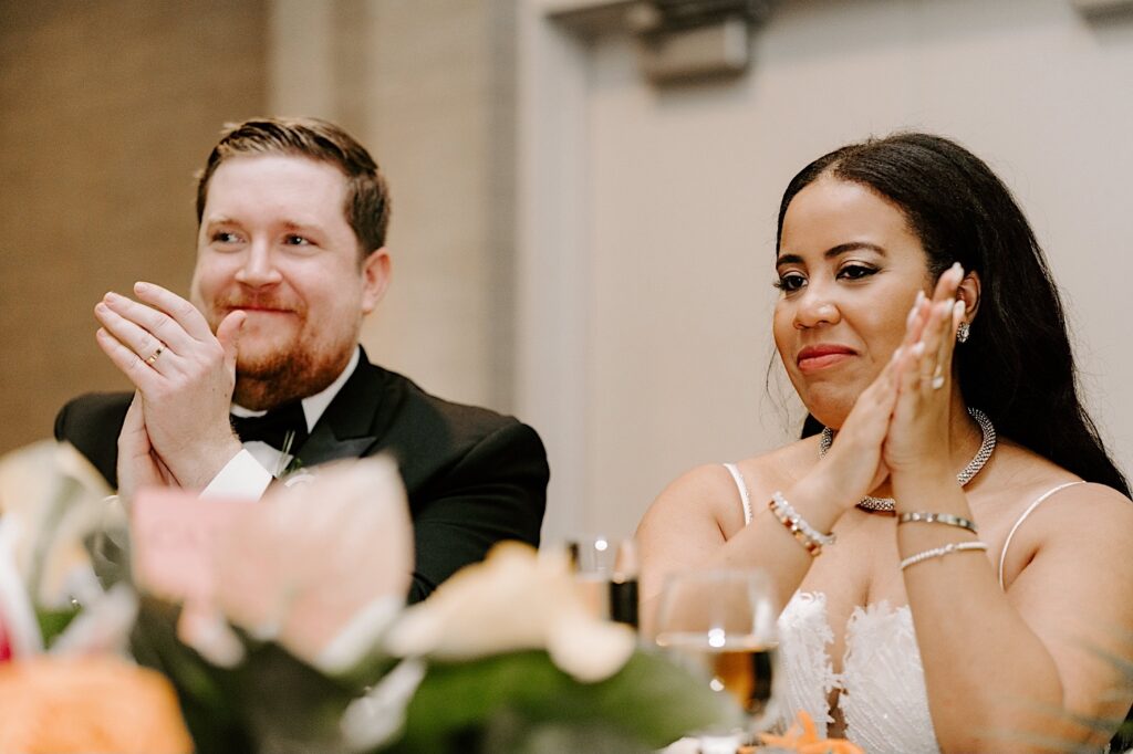 A bride and groom smile while clapping as they sit together at the sweetheart table of their wedding reception