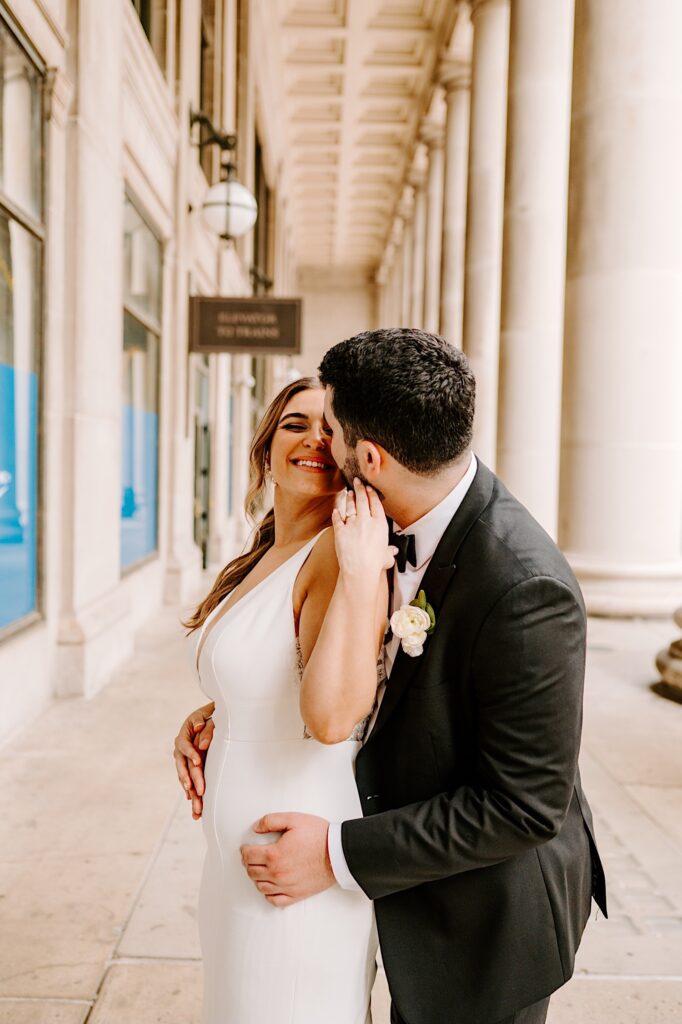 A bride smiles at the groom as he stands behind her and goes in for a kiss while the two are outside of Chicago's Union Station