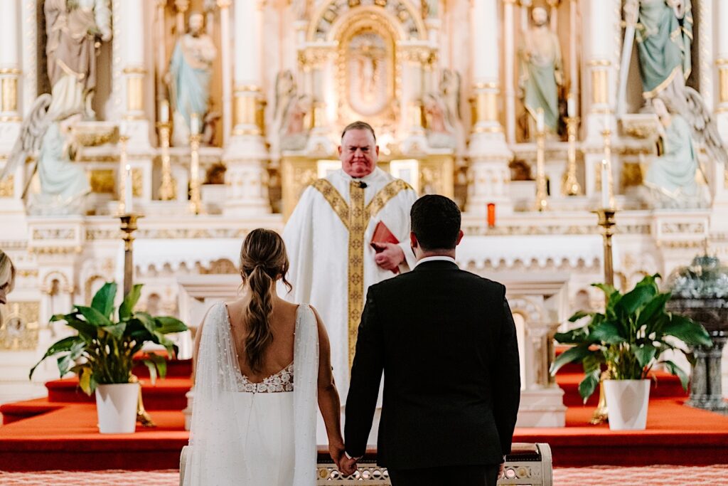 A bride and groom stand next to one another and hold hands while at the altar of St Michael Church in Chicago as the pastor stands in front of them