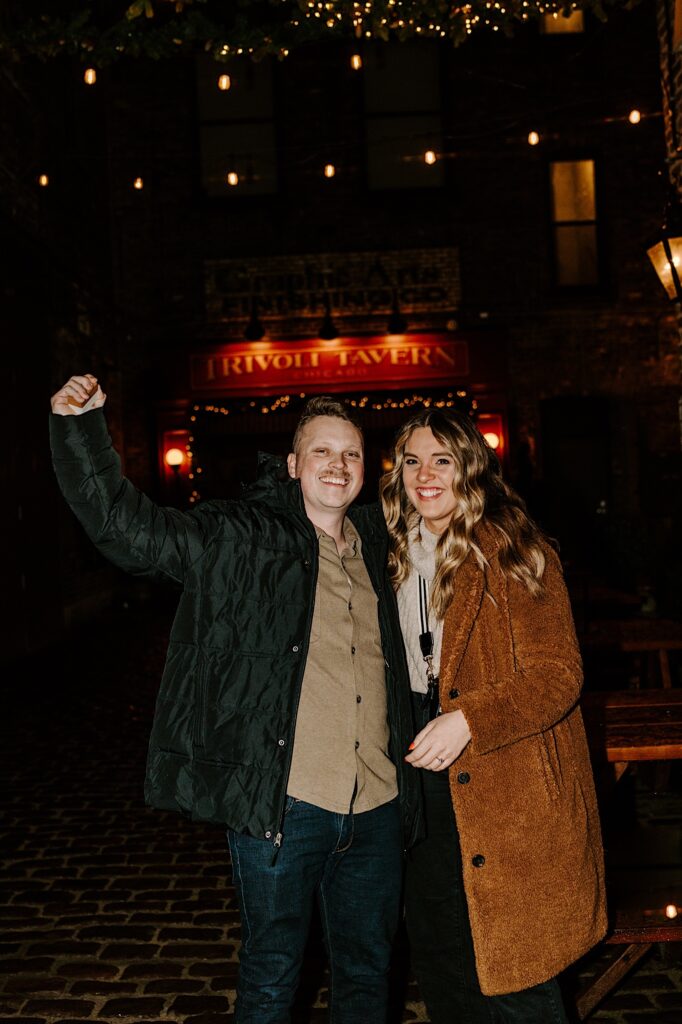 A couple smile at the camera while outside the Trivoli Tavern in Chicago, the man raises his arm in the air as he just proposed to the woman next to him