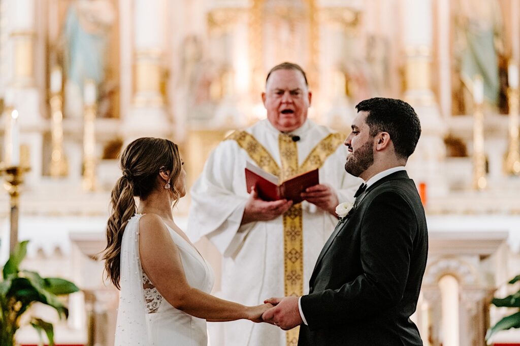 A bride and groom hold hands and face one another while the pastor speaks to them during their wedding ceremony at St Michael Church in Chicago