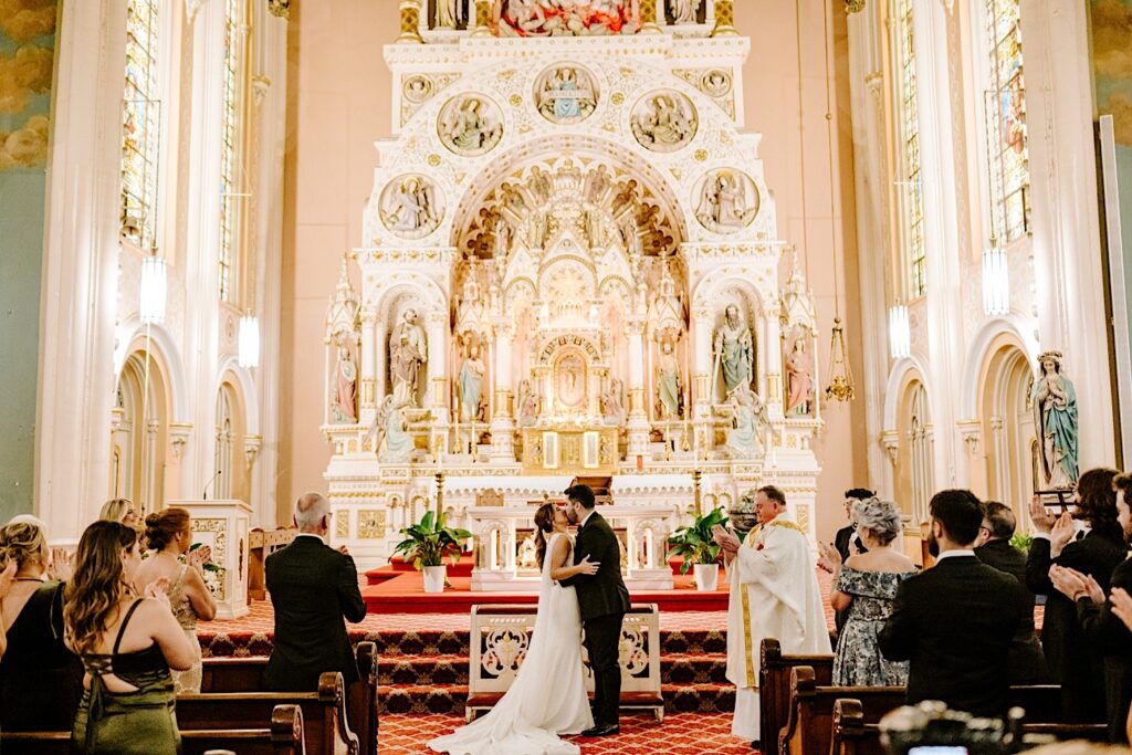 A bride and groom kiss as guests stand and clap during their wedding ceremony in St Michael's Church of Chicago