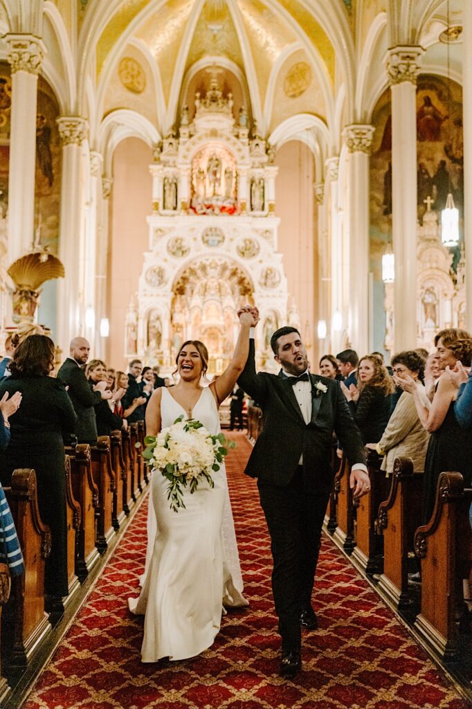 A bride and groom lift their hands in the air as guests around them clap while they walk down the aisle together after their wedding ceremony at St Michael Church in Chicago
