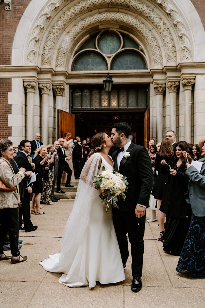 A bride and groom kiss one another while outside of St Michael Church in Chicago as guests around them watch