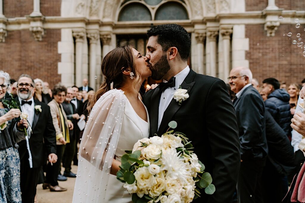 A bride and groom kiss one another while outside of St Michael Church in Chicago as guests blow bubbles around them