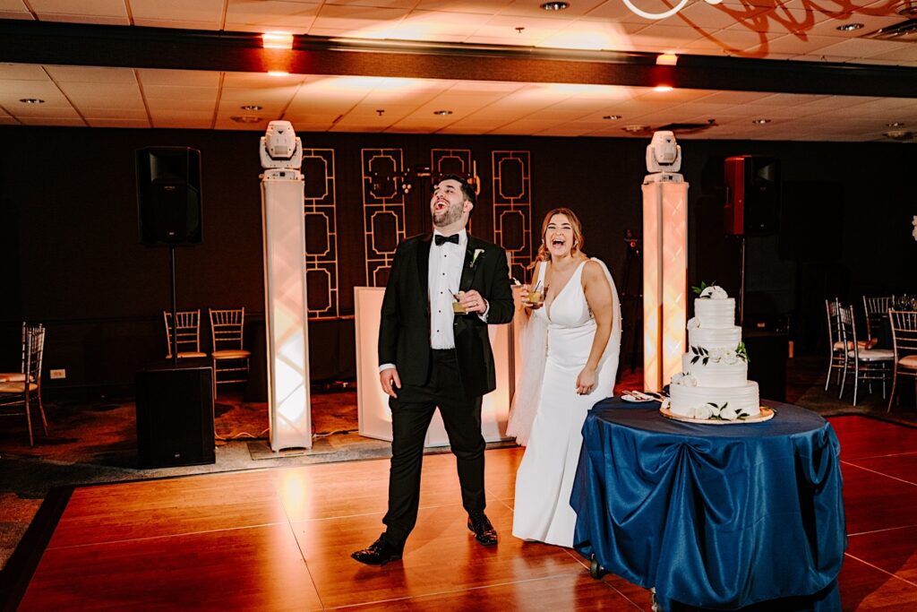A bride and groom laugh while standing next to one another and their wedding cake at the start of their wedding reception inside the Voco Hotel in Chicago