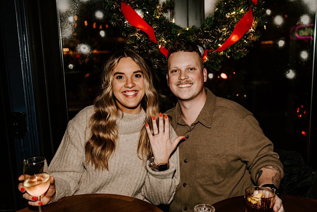 A couple sit inside The Bandit in Chicago and smile at the camera while the woman shows off her engagement ring after their surprise proposal in West Loop