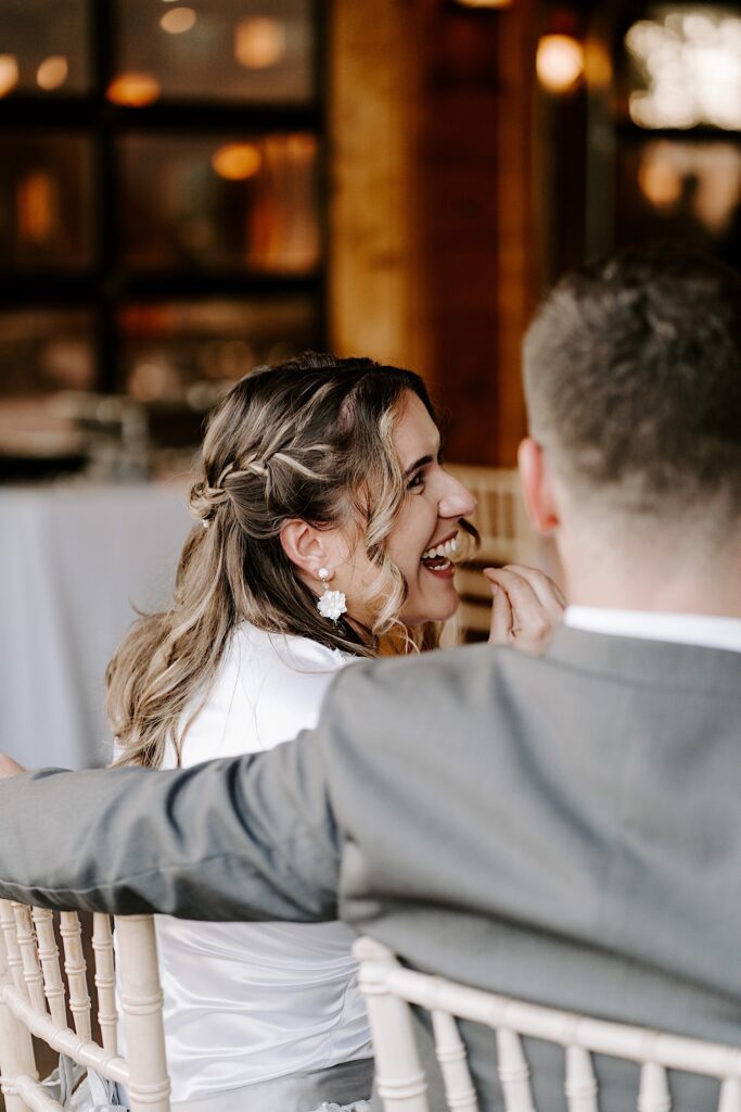 A bride smiles and laughs towards the groom who is sitting next to her and has his arm around the back of her chair