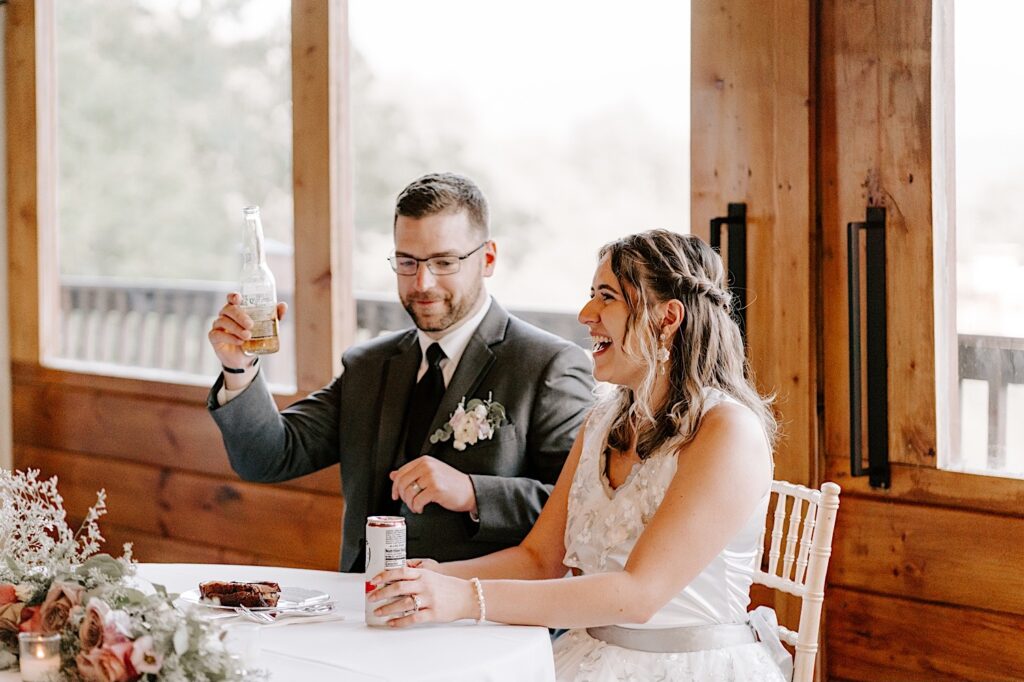 A bride laughs while the groom raises a bottle in the air while the two sit at their sweetheart table