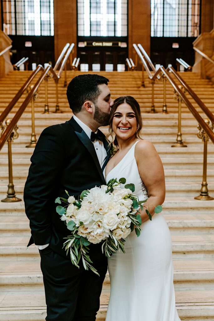 A bride smiles at the camera while the groom kisses her head while the two stand in Chicago's Union Station