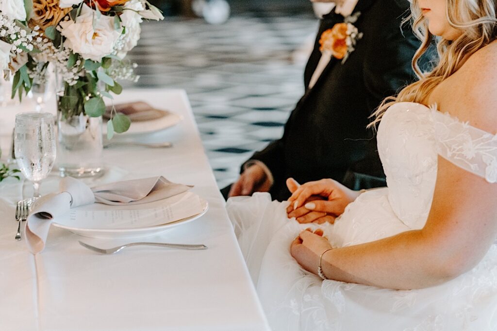 A bride and groom hold hands on the bride's lap while sitting next to one another at their sweetheart table during their wedding reception