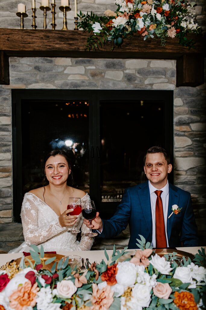 A bride and groom smile at the camera while toasting their glasses together as they sit at their sweetheart table