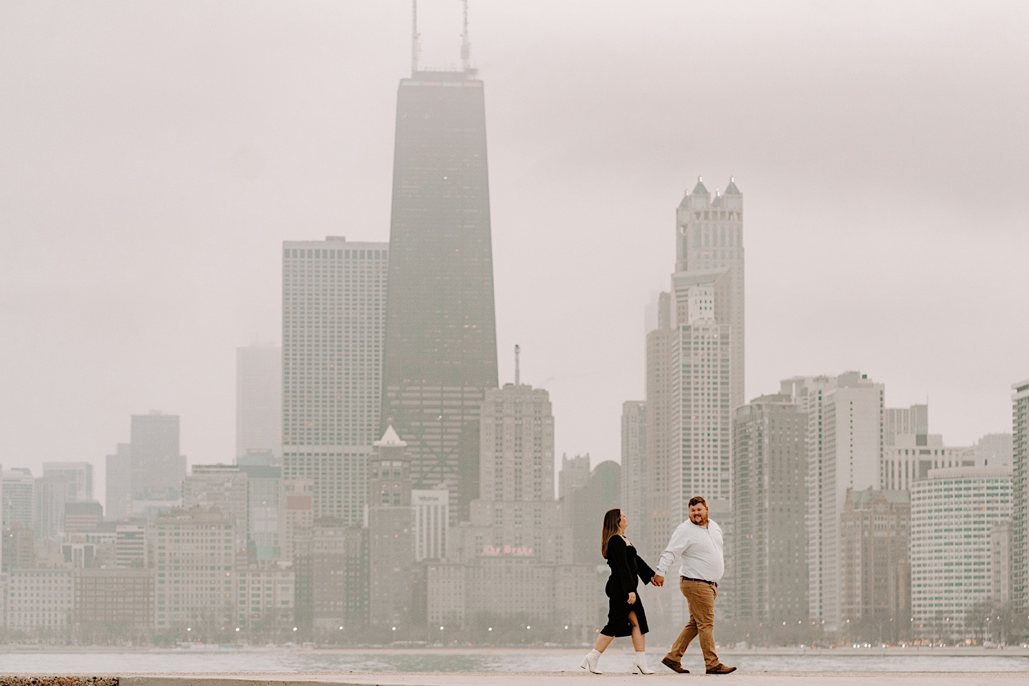 A man looks back towards a woman as the two hold hands and walk along North Avenue Beach in Chicago while taking engagement photos