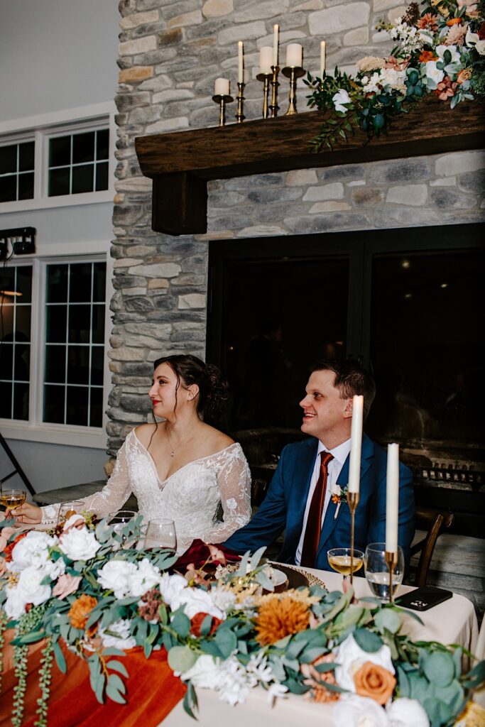 A bride and groom sit together at their sweetheart table and smile to the left at something off camera
