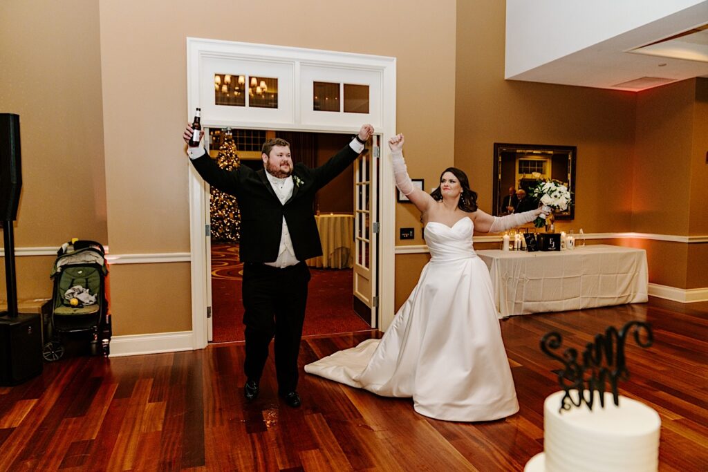 A bride and groom raise their arms as they enter their indoor wedding reception space