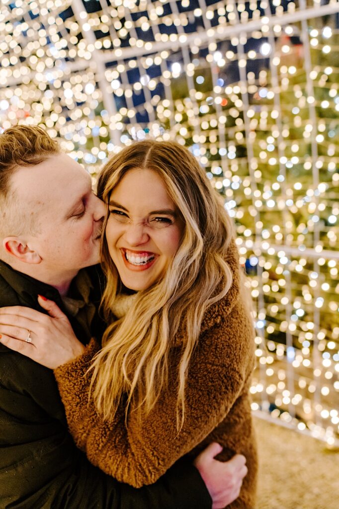 A woman smiles at the camera while being kissed on the cheek by a man while under a tunnel of lights in the Jack Frost Market