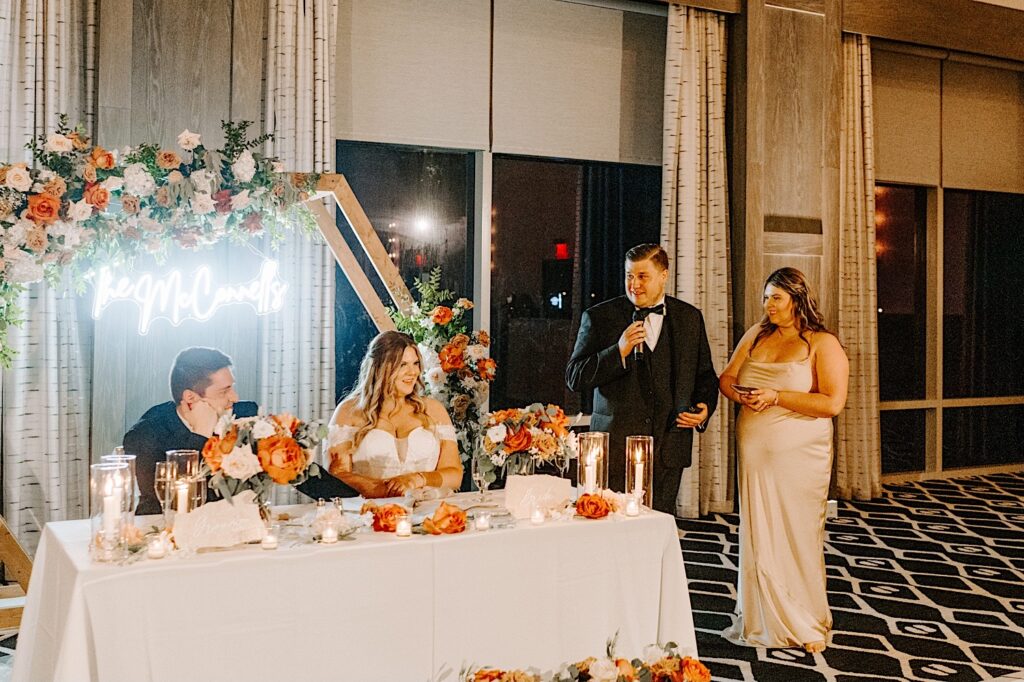 A bride and groom sit at their sweetheart table while a bridesmaid and groomsman stand next to the table and give a speech during their wedding reception